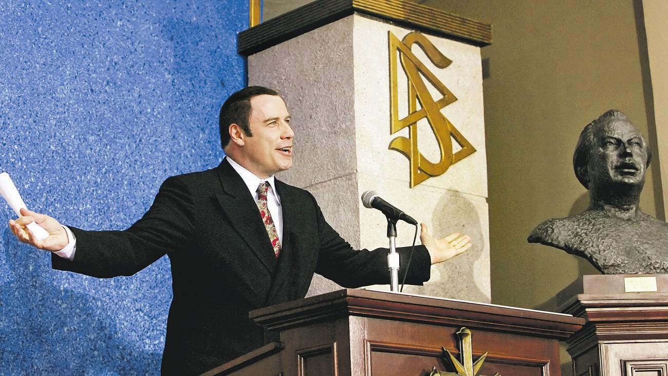 Travolta beszédet tart a szcientológusoknak, de nem beszél róluk (jobbra Hubbard mellszobra)