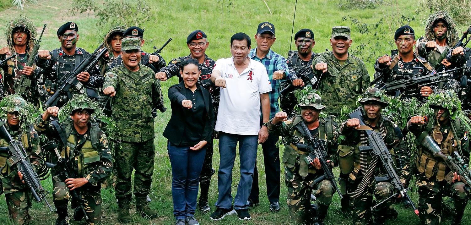 Duterte (középen fehérben) katonák között, szimbólumává vált kézjelével. A keménység népszerűséget hozott neki
