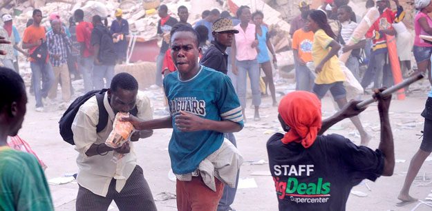Egy férfi beleharap egy élelmiszeres zacskóba, miközben egy áruház tetejéről fosztogatók dobálják le társaiknak a zsákmányt, amelyért az emberek összecsapnak a fővárosban, Port-au-Prince-ben