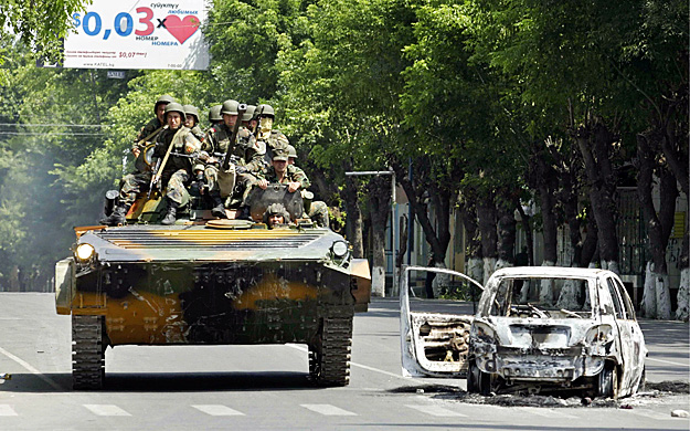 Kiégett autóroncs mellett haladnak el páncélozott harcjárművel kirgiz kormánykatonák Os városában